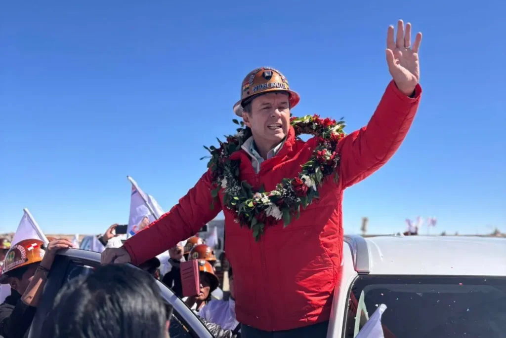 Candidato presidencial boliviano, Jorge "Tuto" Quiroga, saluda desde una camioneta con casco minero que dice ‘Potosí Bolivia’, chaqueta roja y visitiendo una guirnalda floral durante la campaña en Bolivia 2025.