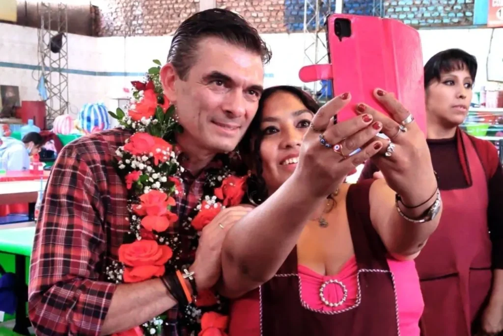 En un mercado techado, el candidato presidencial de Bolivia, Rodrigo Paz con camisa a cuadros y guirnalda de flores rojas se toma una selfie con una simpatizante durante la campaña.